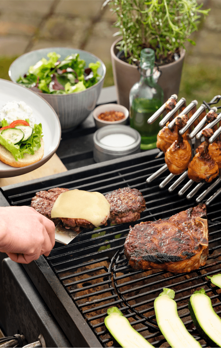 Burger patties, chicken drumsticks on a pot lid holder and chops on the grill.